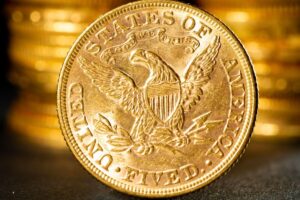 Close-up of a gleaming U.S. $5 gold coin with intricate eagle detailing, set against a background of stacked gold coins. Keywords: gold coin, rare coin auctions, valuable numismatic treasures.