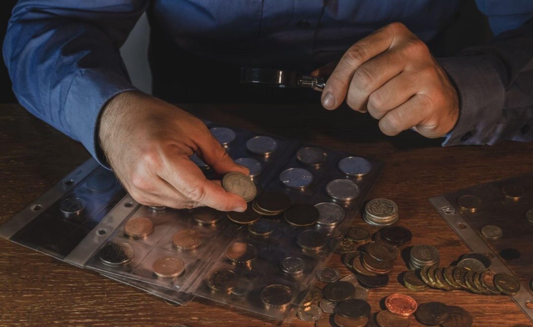 A man carefully examines a rare antique coin with a magnifying glass, surrounded by a collection of historical coins displayed in protective sleeves. Keywords: rare coin, antique coin auctions, coin examination.