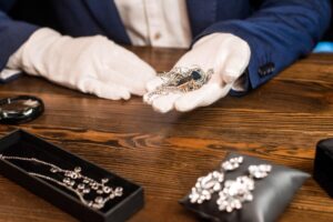 A jewelry appraiser wearing white gloves carefully holding a vintage necklace, surrounded by antique brooches and necklaces on a wooden table. Keywords: vintage jewelry appraisal, antique jewelry evaluation, expert jewelry appraisal.