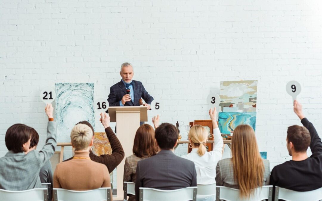 An auctioneer leading a live art auction with bidders raising paddles for valuable paintings. A competitive bidding environment driving artwork prices higher. Perfect for those looking to sell artwork at auction